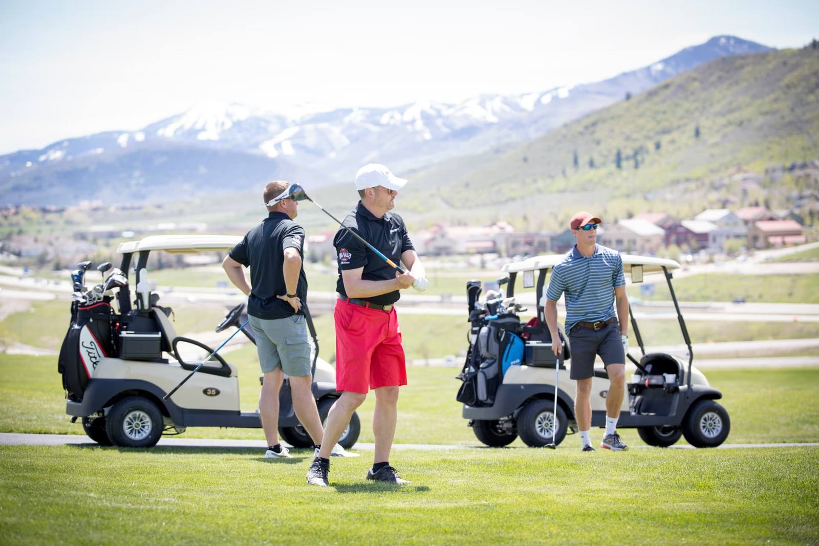 Three golfers on a lush Utah course with snow-capped mountains and golf carts nearby