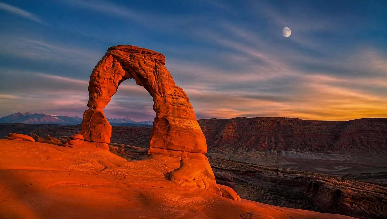 Delicate Arch in Arches National Park at sunset with a visible moon in the sky