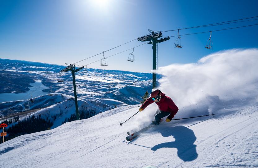 A skier carving fresh powder on a sunny Utah mountain with chairlifts and a lake valley below