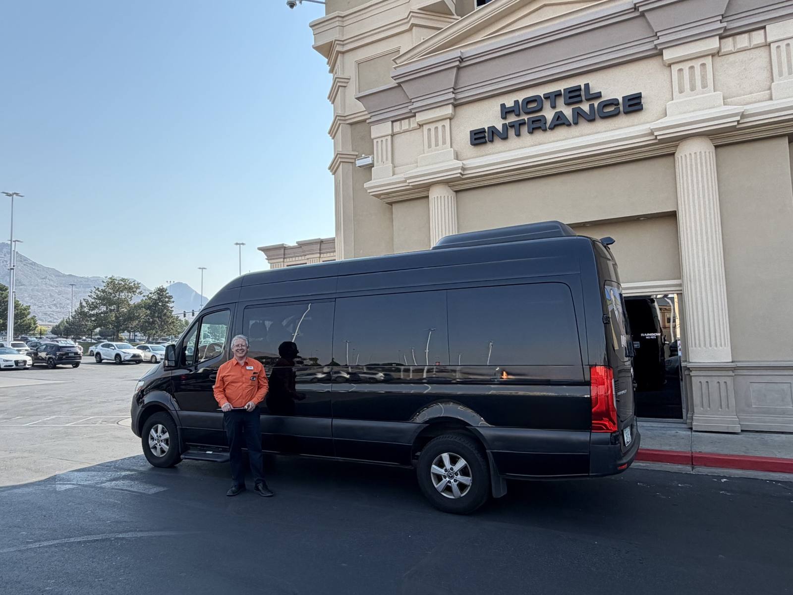 Chauffeur standing beside a black Mercedes Sprinter van at a hotel entrance